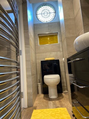 Un baño pequeño con inodoro, un ventanal con un diseño decorativo de vidrio y un tapete amarillo en el suelo.