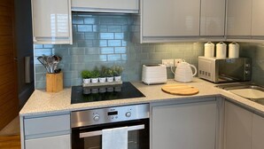 Een moderne keuken met grijze kastjes, een ingebouwde oven, een magnetron en een broodrooster. Op het aanrecht staan potplanten en er ligt een houten snijplank.