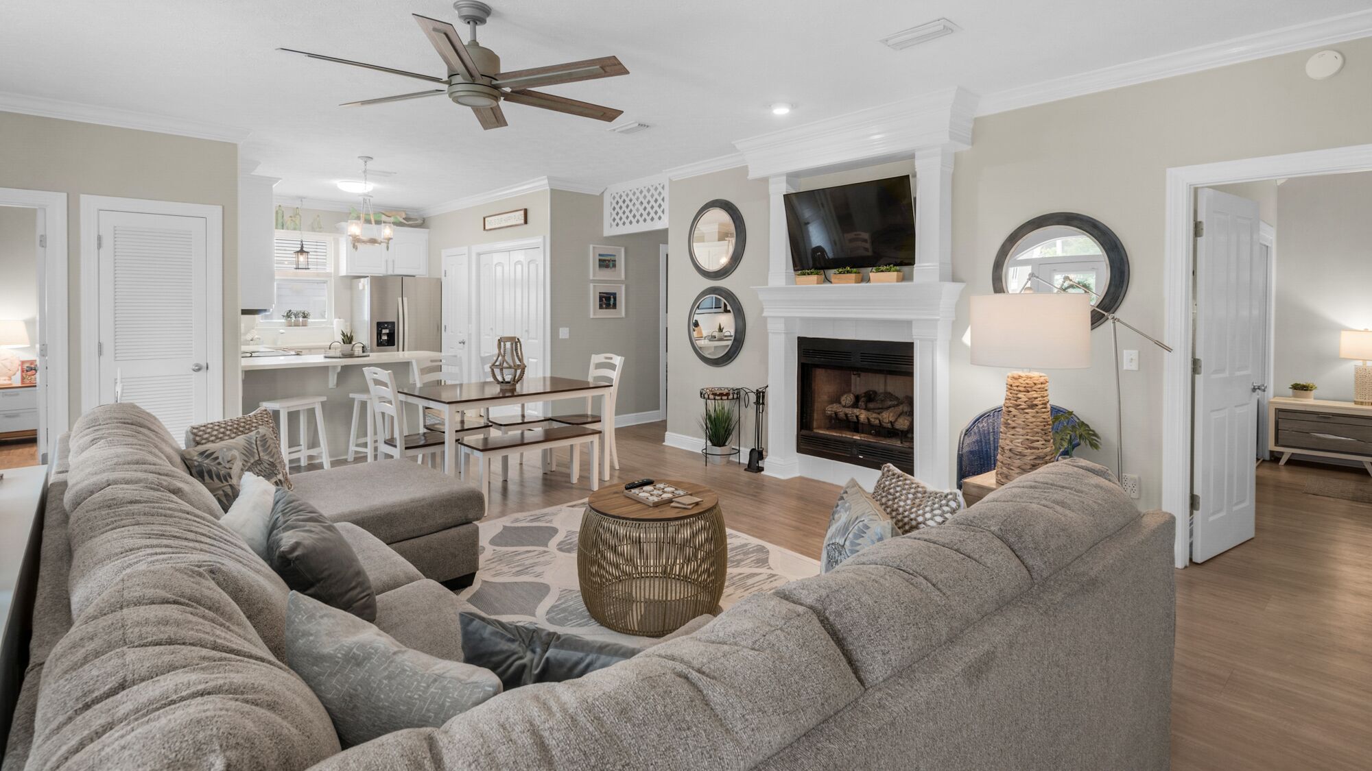 A spacious living room with a large gray sectional sofa, a dining area with a white table and chairs, a fireplace, and a kitchen in the background.
