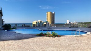 A swimming pool with a circular design, surrounded by a paved area with lounge chairs and a building with a balcony in the background.