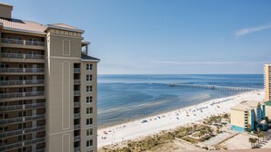 A beachfront hotel with multiple floors, a pier extending into the ocean, and a sandy beach with umbrellas and beach chairs.