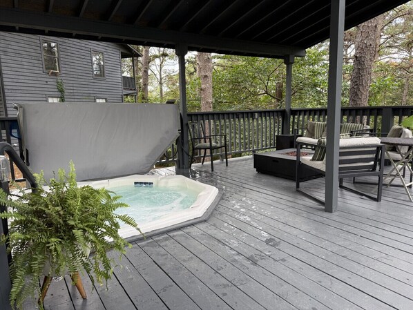 A covered outdoor deck with a hot tub, seating area, and a view of a house.