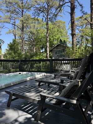 A wooden deck with lounge chairs, a hot tub, and a cabin in a forested area.