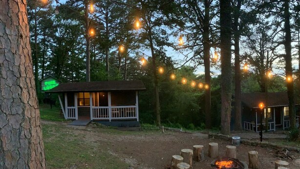 A rustic cabin with a porch, surrounded by trees and a fire pit.