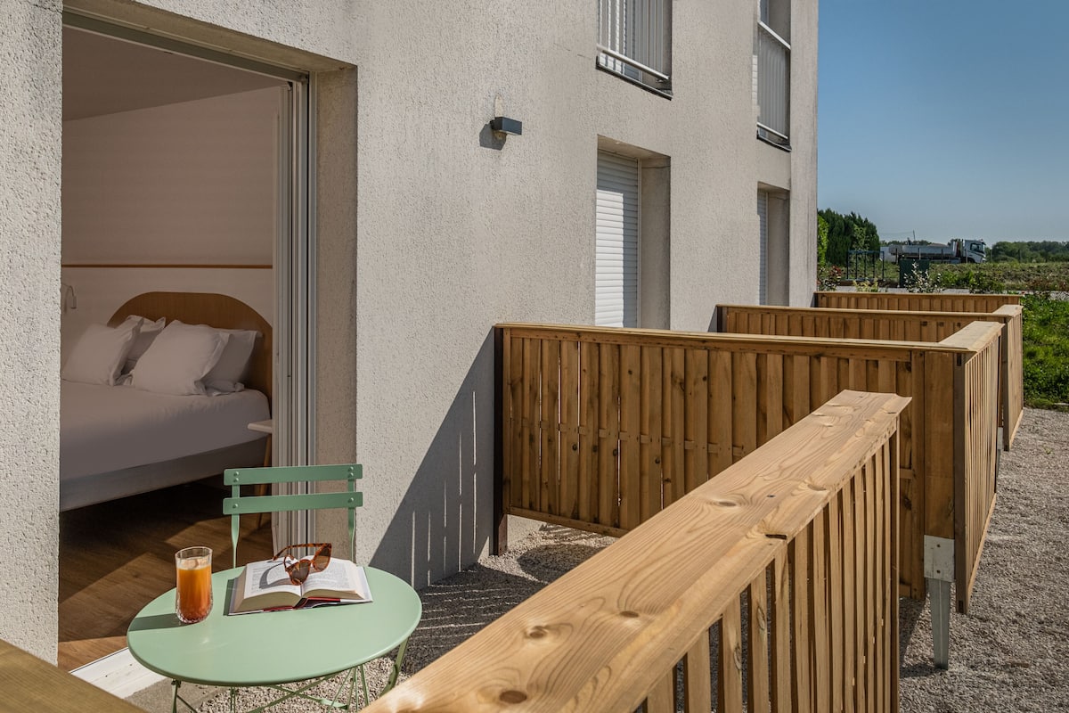 A room with a bed, a table with a drink and book, and a view of the outside.