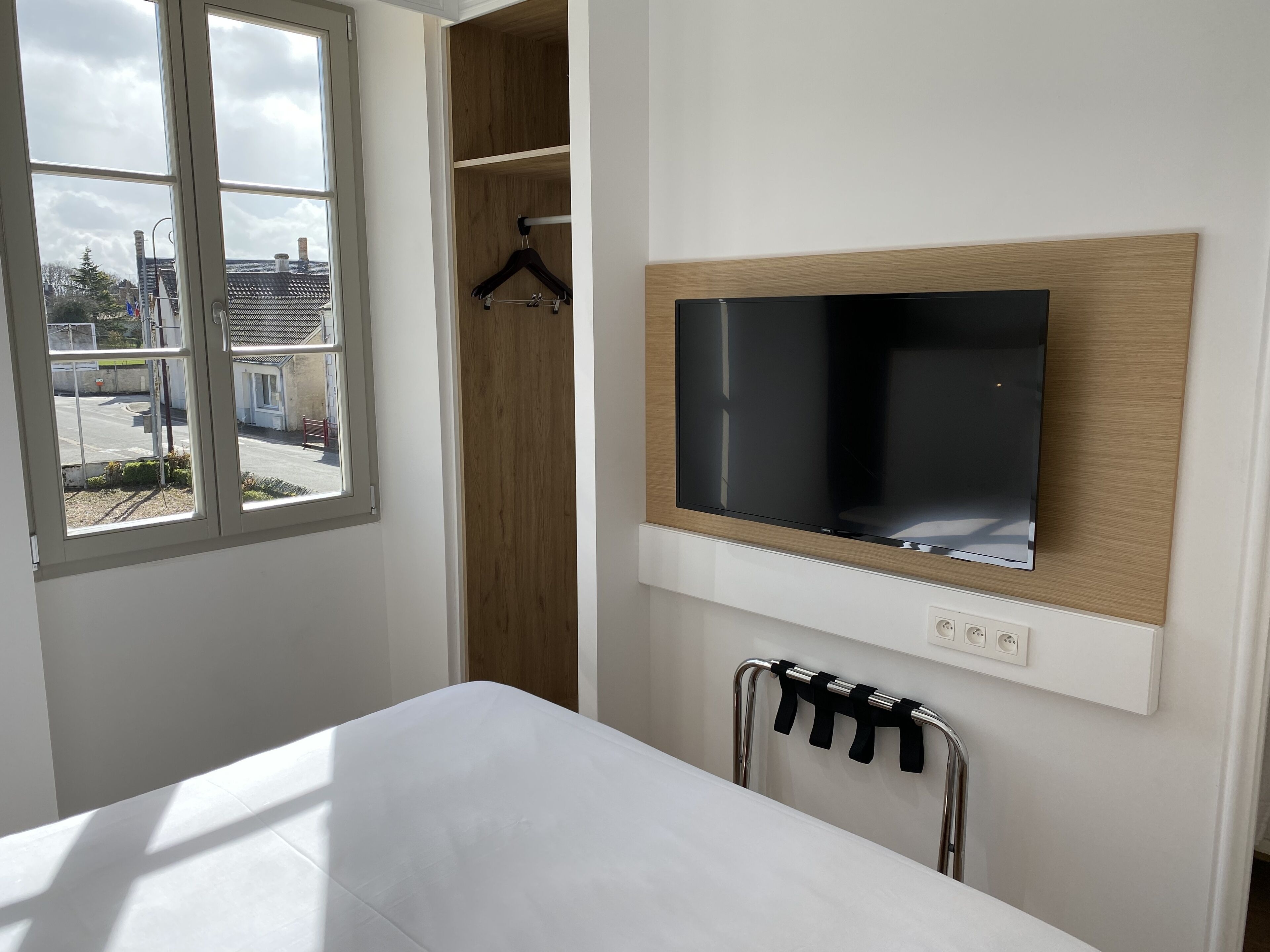 A modern hotel room with a bed, a TV mounted on the wall, a wooden wardrobe, and a view of the outside.