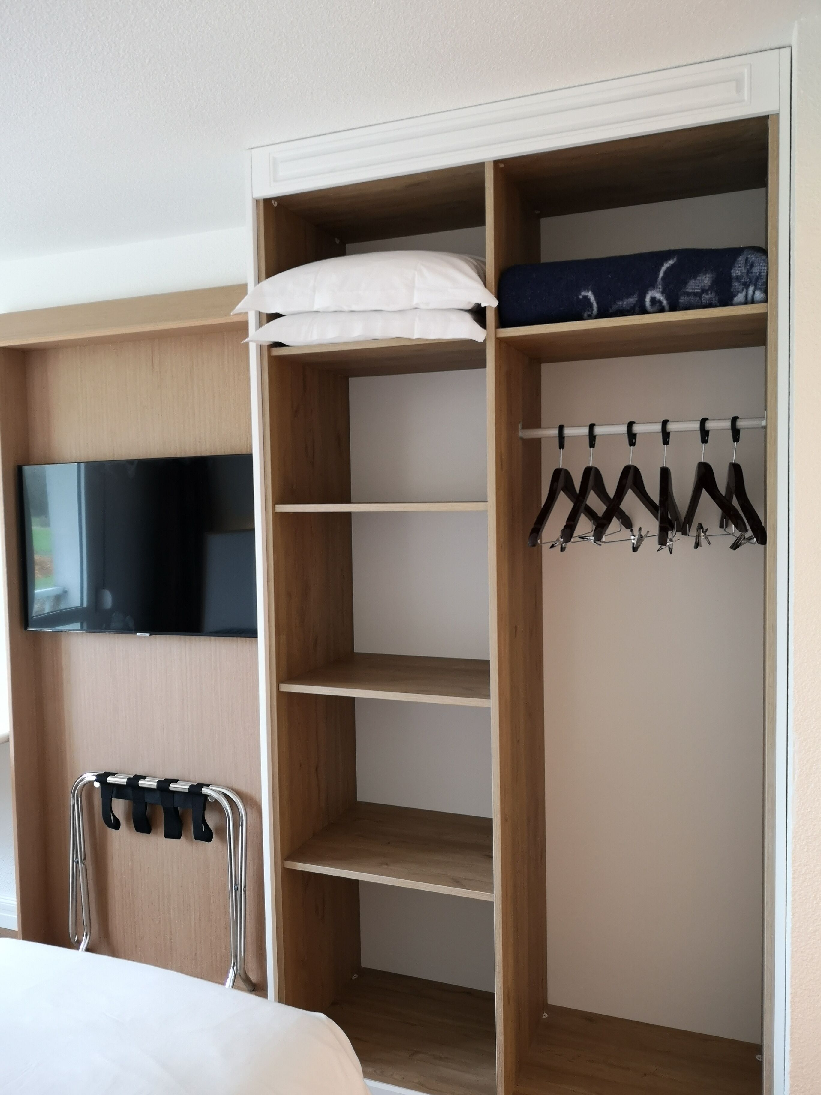 Un dormitorio con cama, televisor montado en la pared, armario de madera con estantes y perchero para ropa.