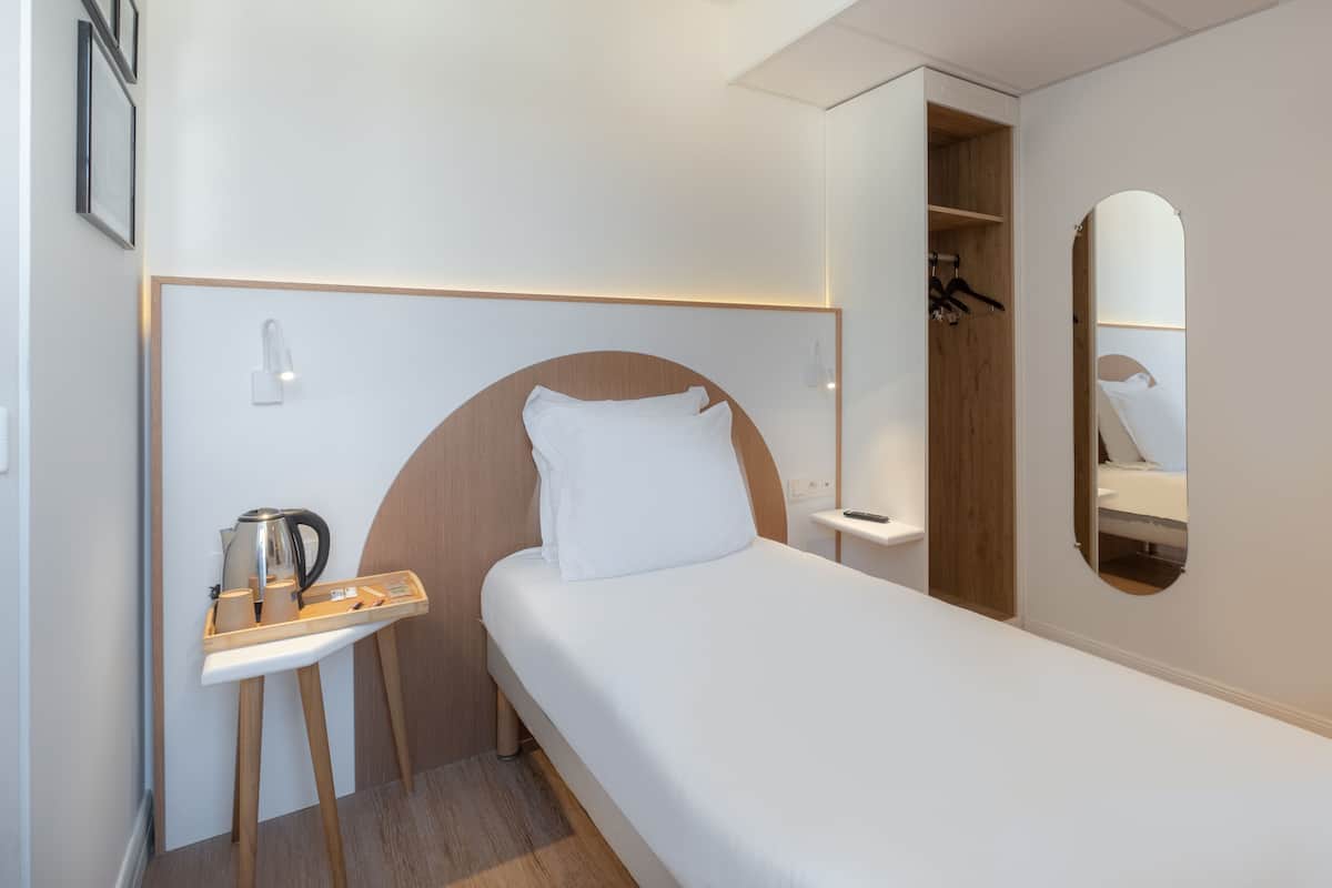 A compact hotel room with a single bed, a small bedside table with a kettle, a mirror, and a wardrobe.