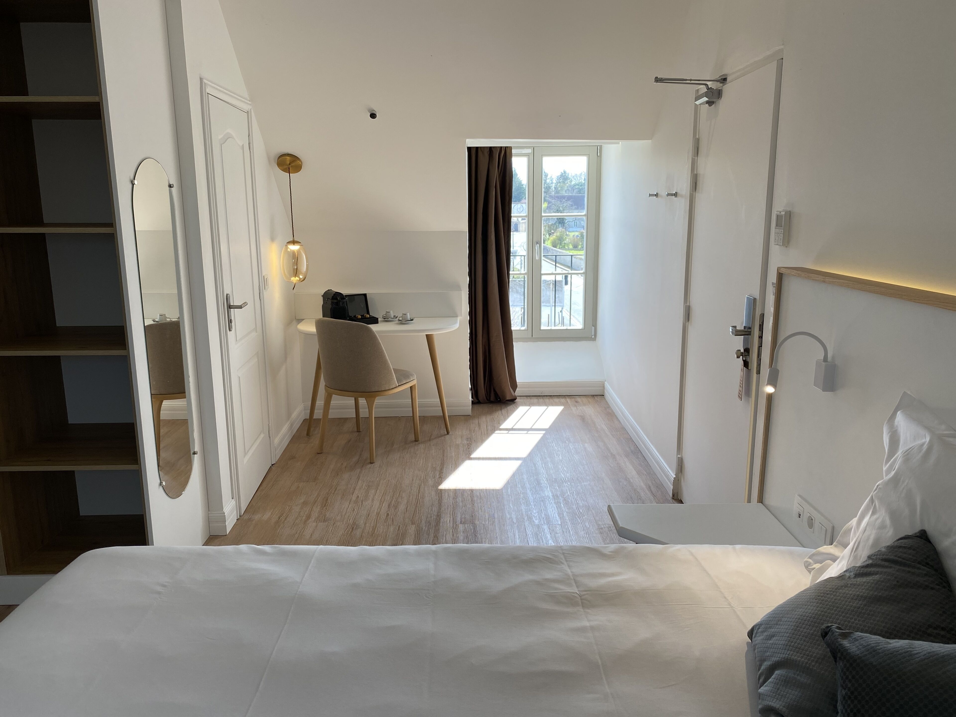 A hotel room with a bed, a desk, and a chair. There is a window with curtains, a door, and a mirror.