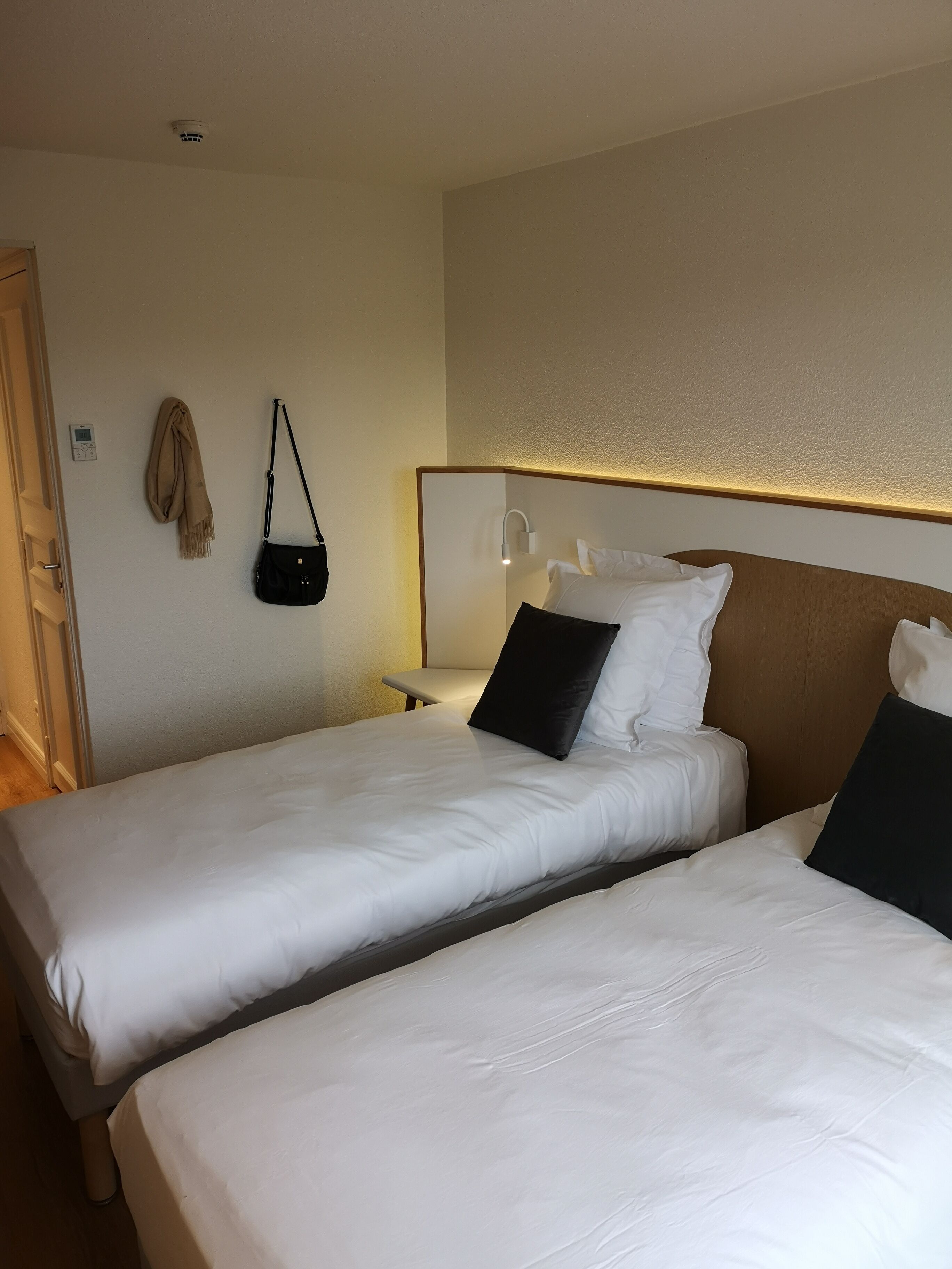 A hotel room with two single beds, a bedside table with a lamp, a wall-mounted heater, and a hanging bag.