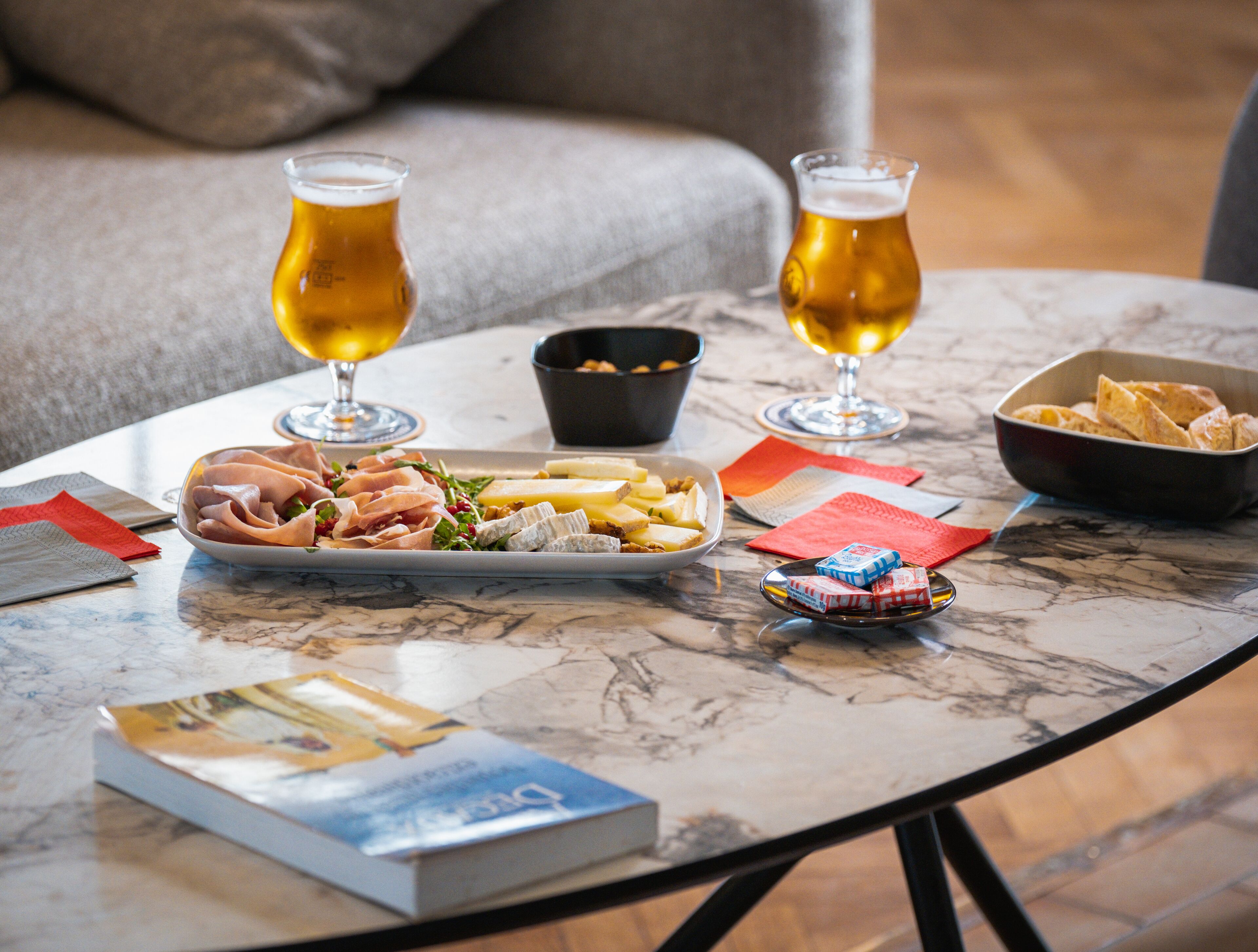 Una mesa con un libro, un plato de comida, dos vasos de cerveza y un bol de patatas fritas.