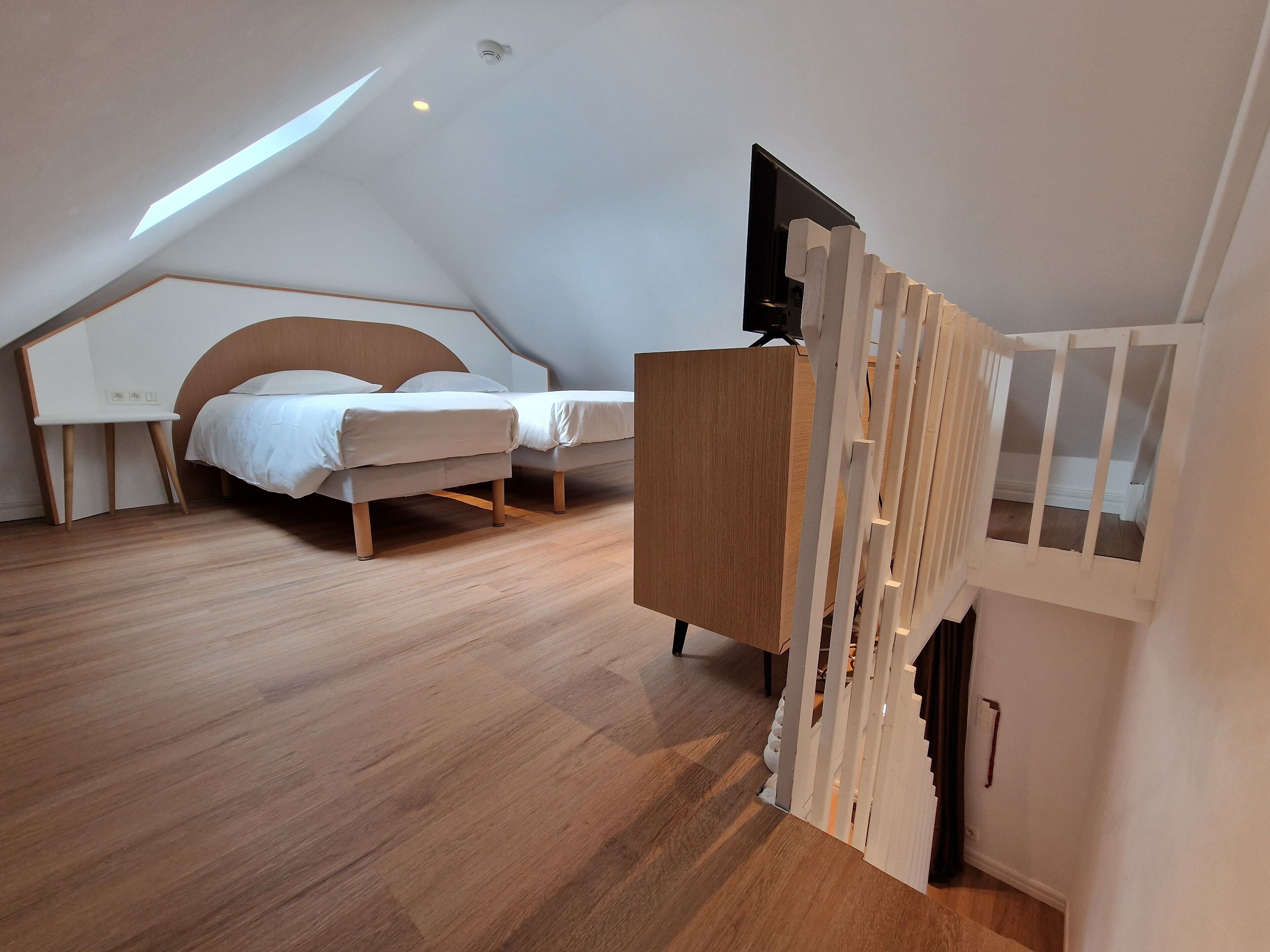 A modern bedroom with a sloped ceiling, a wooden bed, a white desk, and a television mounted on a white partition.
