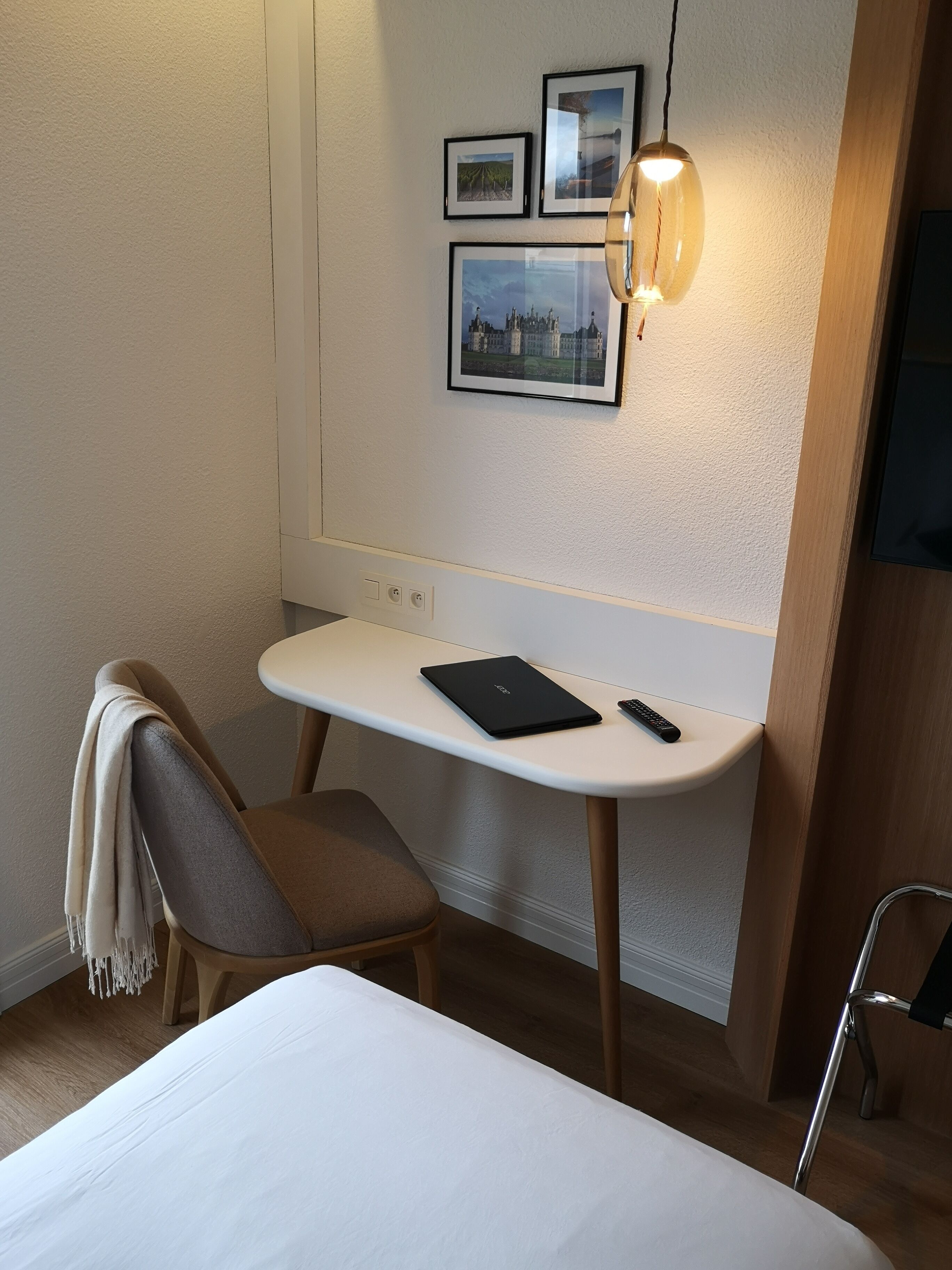 Habitación de hotel con cama, escritorio con una computadora portátil y control remoto, silla con una manta, y cuadros en la pared.
