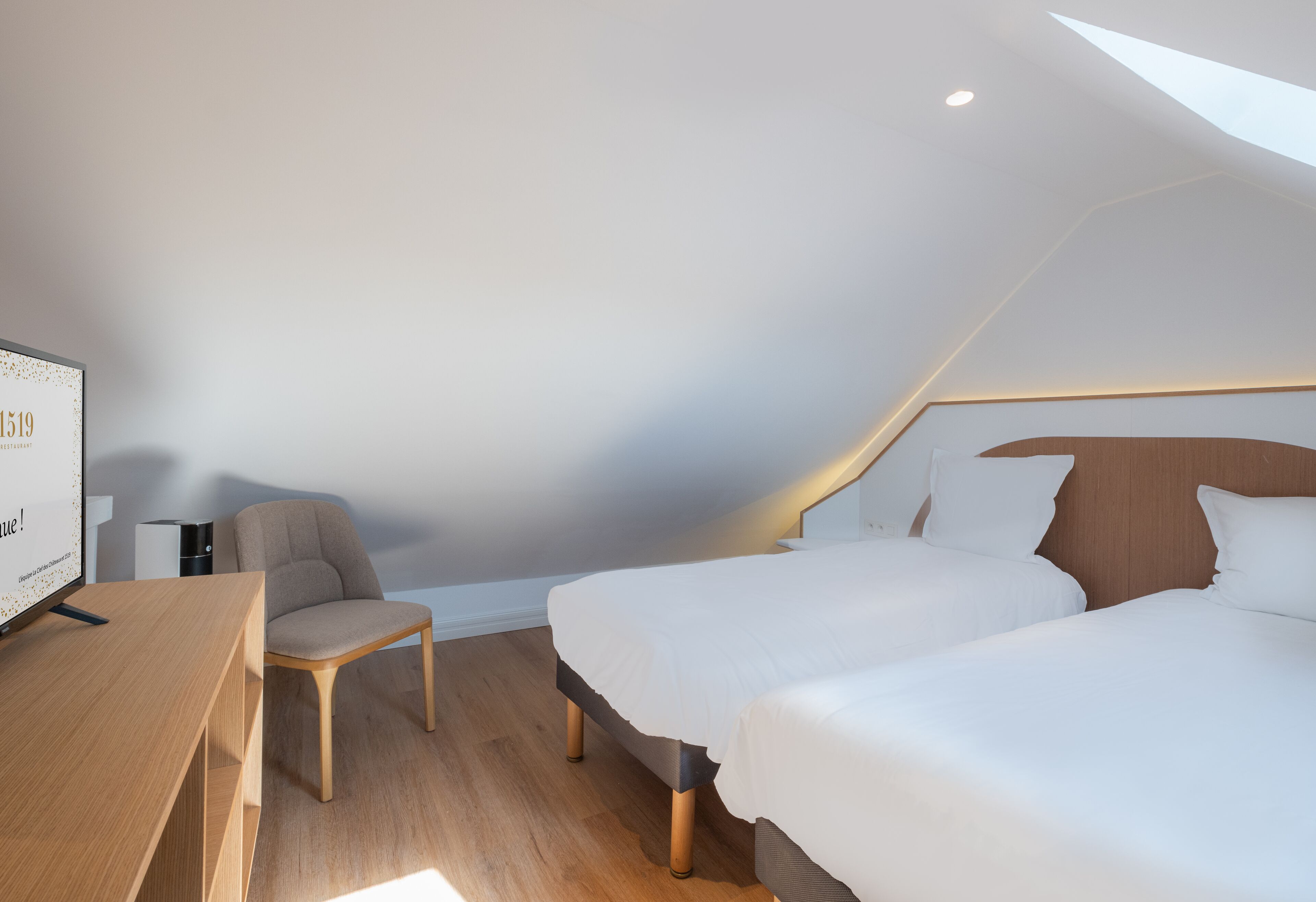 A modern hotel room with a wooden desk, two beds, a chair, and a ceiling light.
