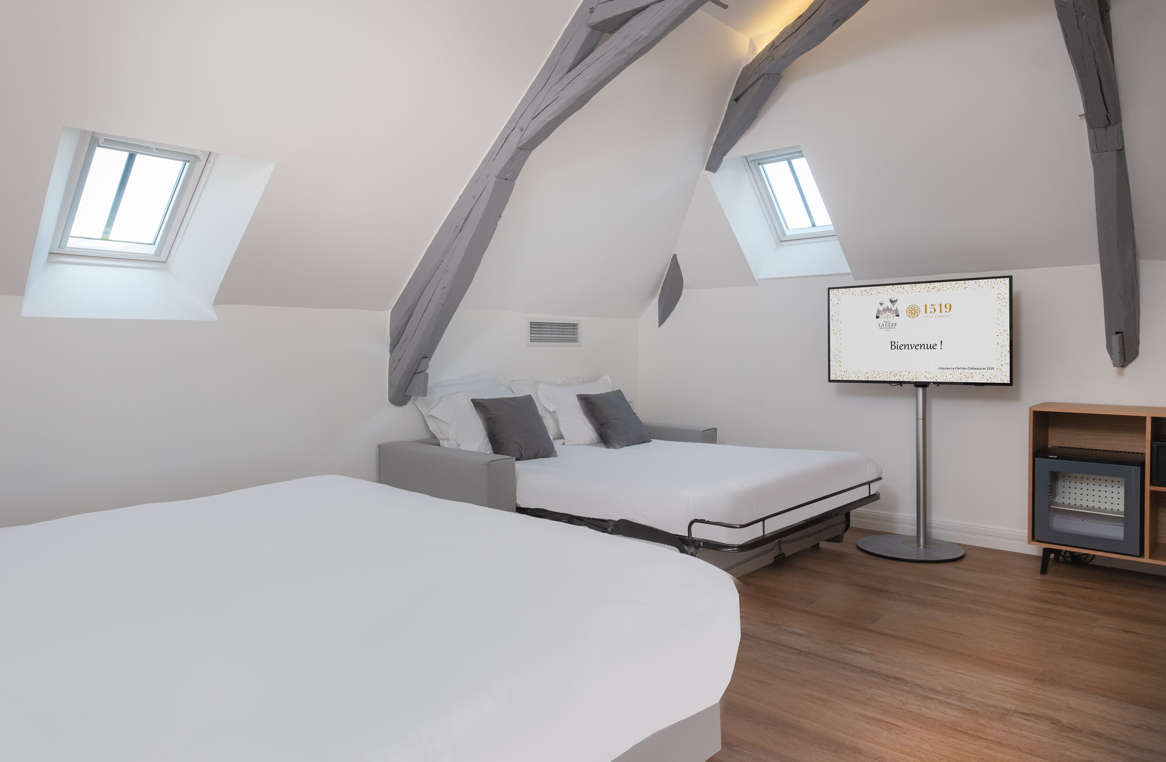 Ein modernes, helles Zimmer mit schrägedachähnlicher Decke, einem Bett, einem Schreibtisch und einem Fernseher, der eine Willkommensnachricht anzeigt.