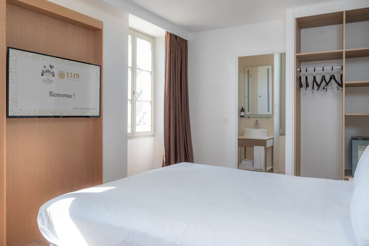 A hotel room with a large bed, a wooden wardrobe, a bathroom with a sink and mirror, and a framed welcome sign on the wall.