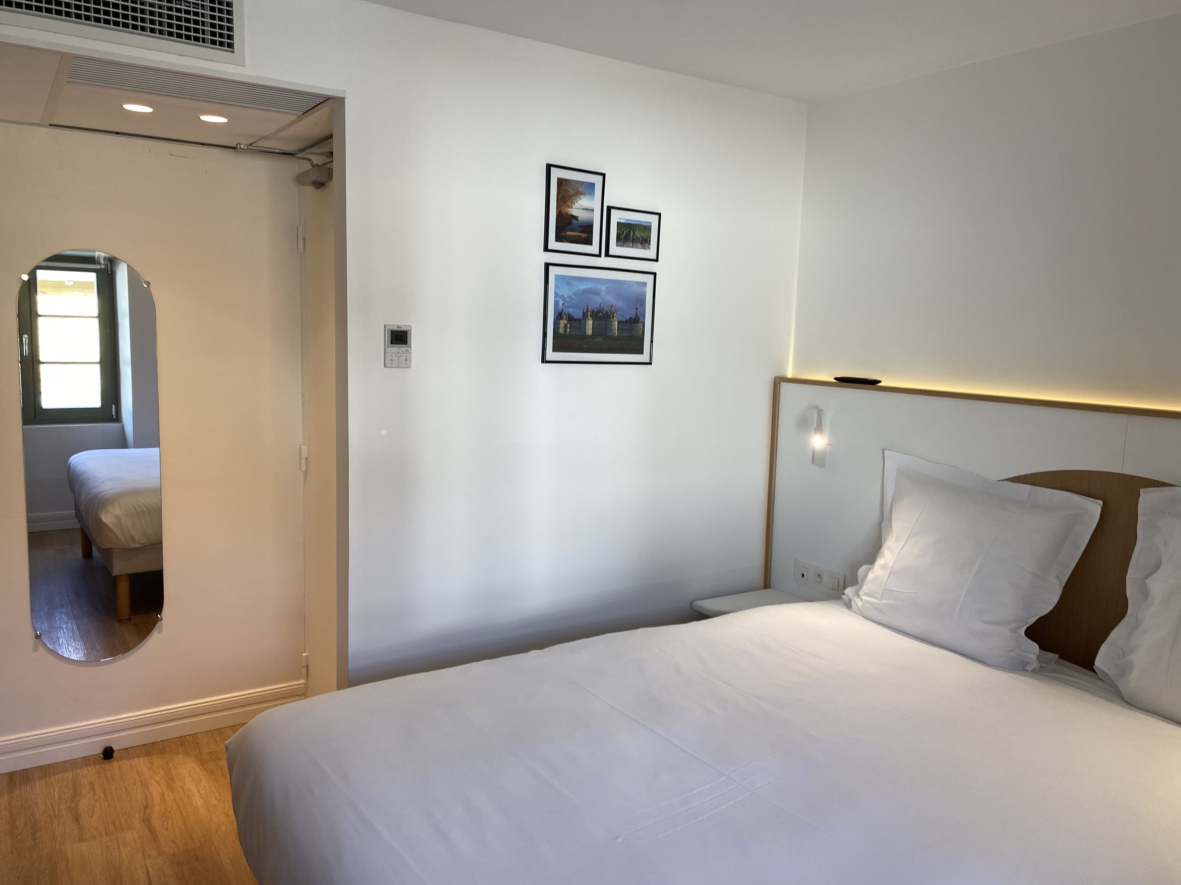 A hotel room with a bed, a bedside table, a mirror, and two framed pictures on the wall.