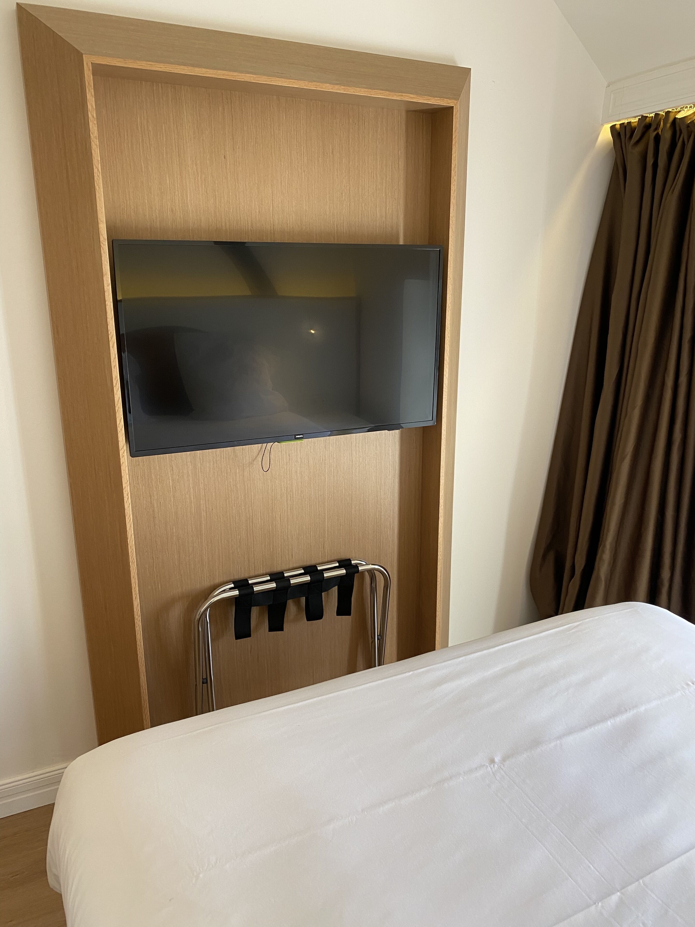 A hotel room with a bed, a wooden wardrobe, a TV mounted on the wardrobe, and a towel rack.