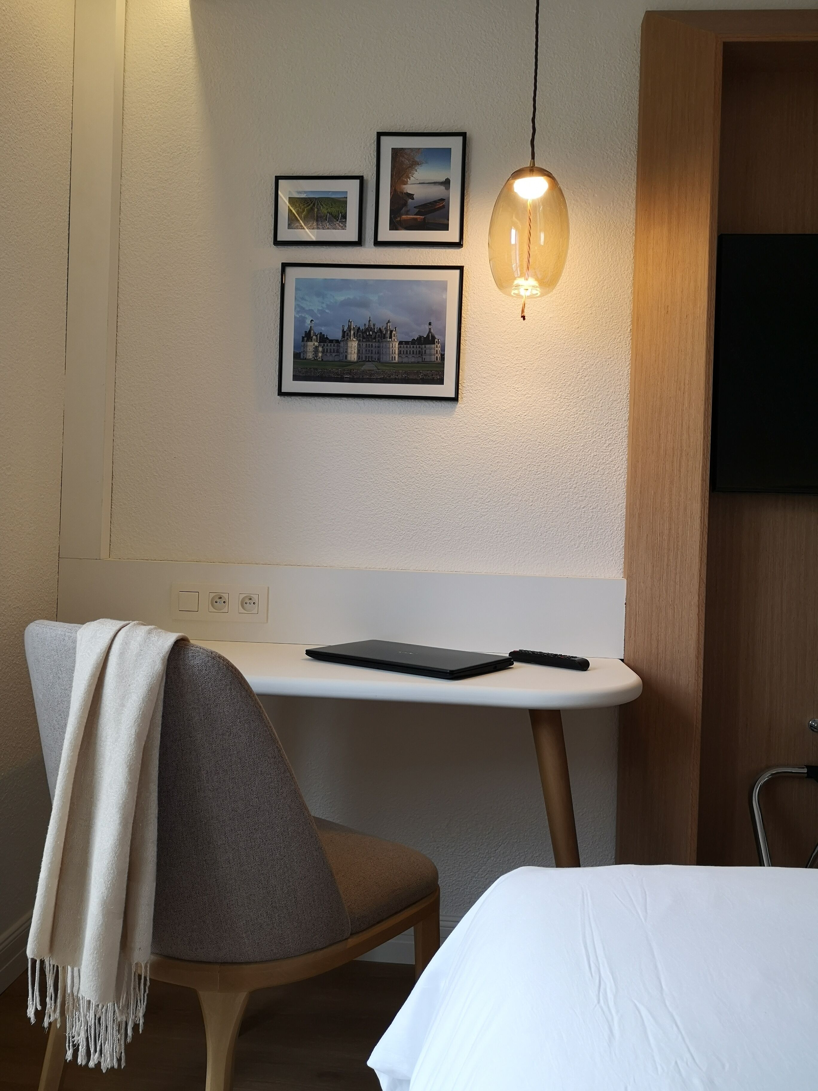Habitación de hotel con cama, una silla con manta, una mesita con control remoto y cuadros en la pared.