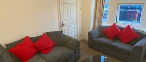 A living room with two grey sofas, each with red cushions, a black coffee table, a white door, and a window with curtains.