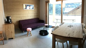 Una habitación acogedora con pared de madera, un sofá púrpura, una mesa de vidrio y una mesa de comedor de madera con sillas.