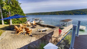 A lakeside patio with Adirondack chairs, a fire pit, and a wooden deck overlooking the water. - Just Bluffin': Keuka Lake Luxury Rental + Hot Tub! Close to Wineries (Penn Yan)