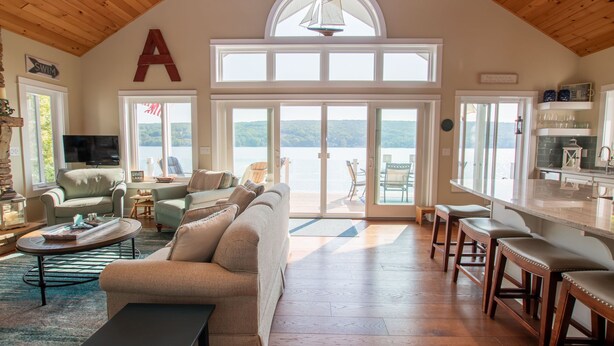 A spacious living area with a large window overlooking a body of water, a cozy seating arrangement, and a kitchen area with bar stools. - Just Bluffin': Keuka Lake Luxury Rental + Hot Tub! Close to Wineries (Penn Yan)