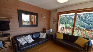 Una sala de estar con piso de madera, un sofá de cuero oscuro, un televisor y un ventanal con vistas a una montaña boscosa.