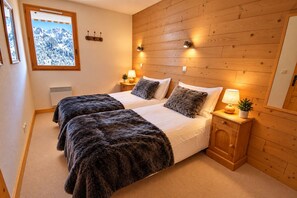 A bedroom with two beds, wooden paneling on the walls, bedside lamps, and a window with a view of snow-covered mountains.