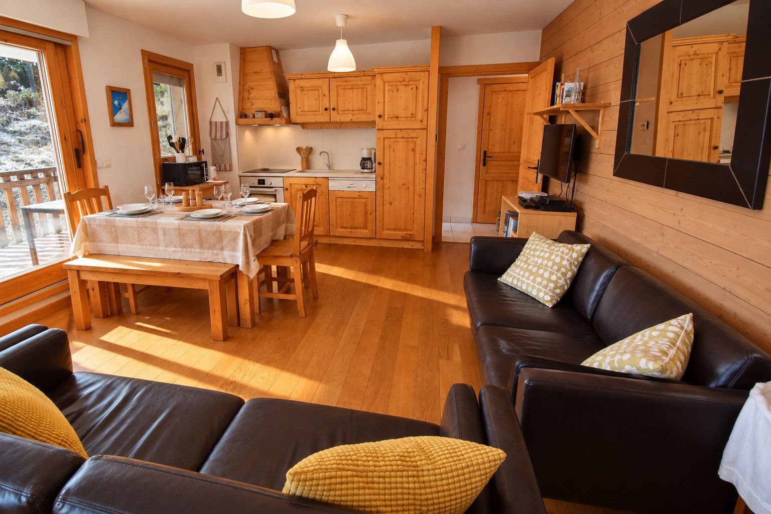 A cozy living room with a black leather sofa, wooden dining table, and a kitchen area with wooden cabinets and a large mirror.
