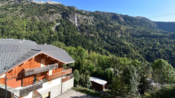 Una cabaña de madera con balcón, rodeada de un paisaje montañoso cubierto de bosque.