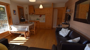 Una sala de estar acogedora con un sofá de cuero negro, una mesa de comedor de madera con sillas y una cocina con alacenas de madera y un colgador para ollas.