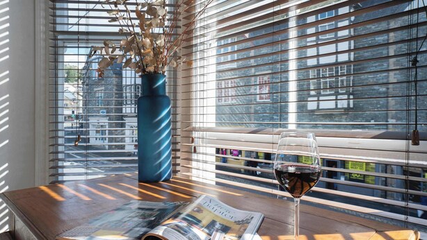 Ein Holztisch mit einer Zeitung, einem Glas Rotwein und einer Vase mit Trockenblumen neben einem Fenster mit Jalousien.