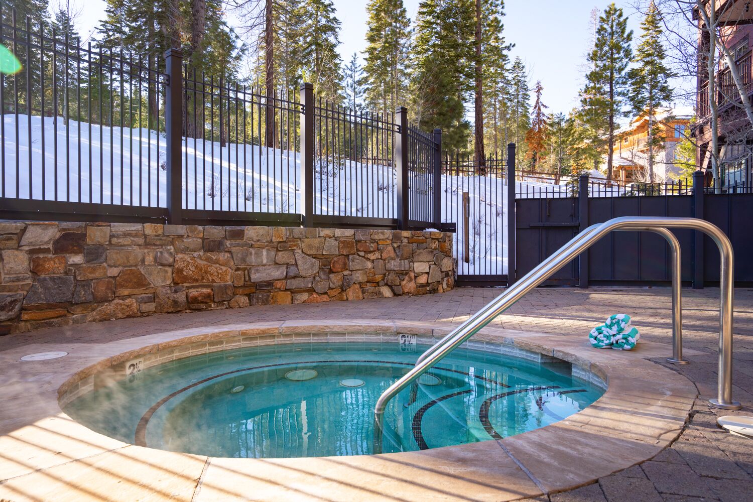 A hot tub surrounded by stone walls and a metal fence, with a snowy landscape and trees in the background.