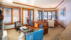 A spacious living room with a large brown leather sofa, blue armchairs, a dining table with chairs, and large windows with wooden blinds.