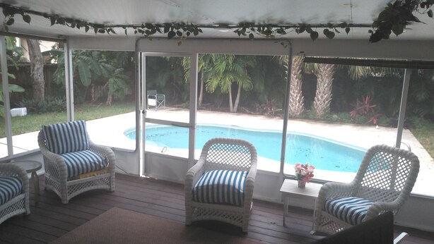 A covered patio with wicker chairs, a small table, and a view of a swimming pool. - Comfortable 3 bedroom 3 bath beach house .pool..3 minute walk to beach. (Vero Beach)