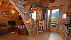 A rustic wooden cabin interior with a fireplace, wooden dining table, chairs, and a kitchen area. - Les Cimes - Five Bedroom Apartment, Sleeps 10 (Vaujany)