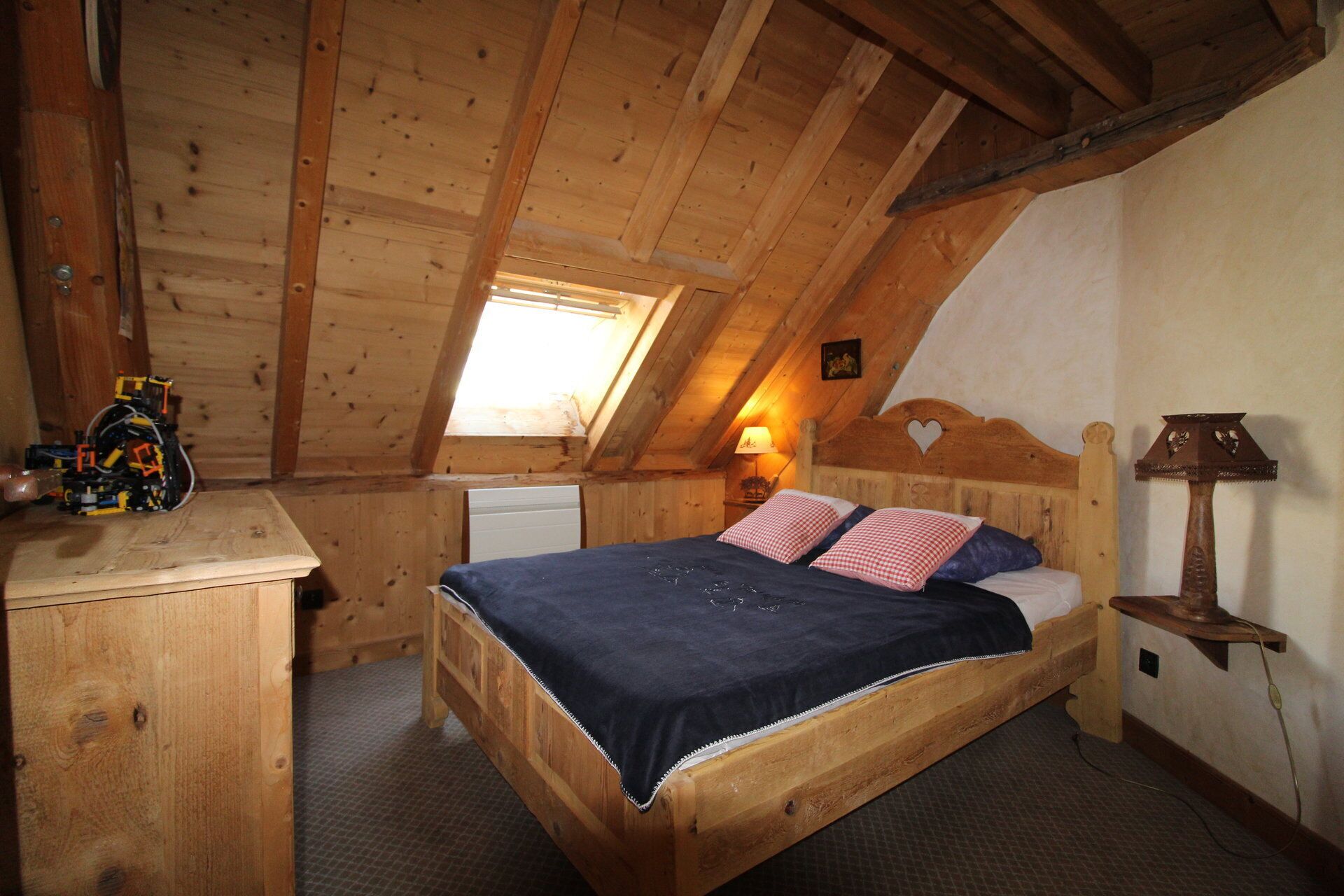 Una habitación de madera con techo inclinado, una cama con colcha de cuadros, un escritorio con lámpara y un ventanal pequeño.