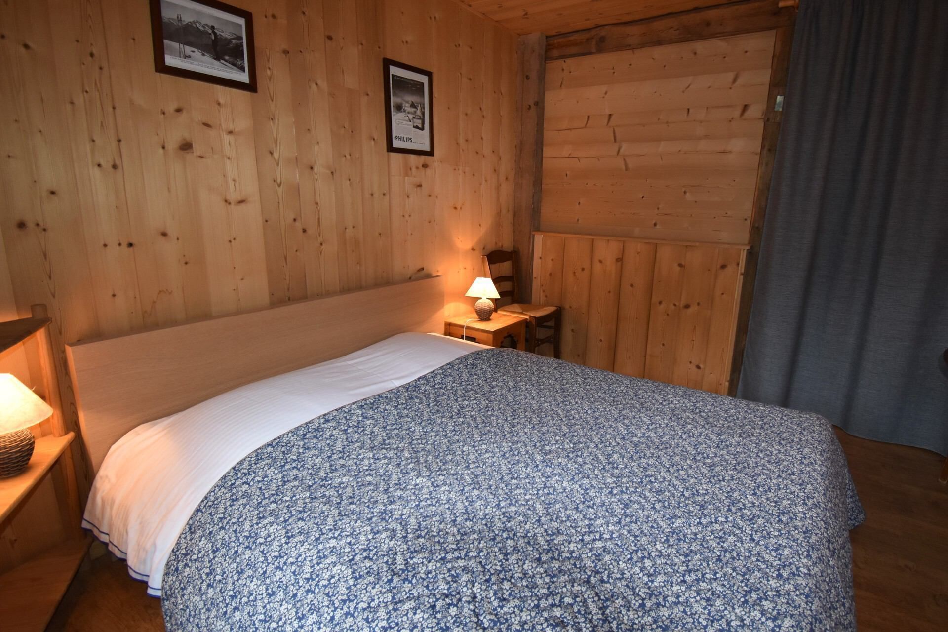 Quarto com painéis de madeira, uma cama, um criado-mudo com abajur e duas molduras na parede.