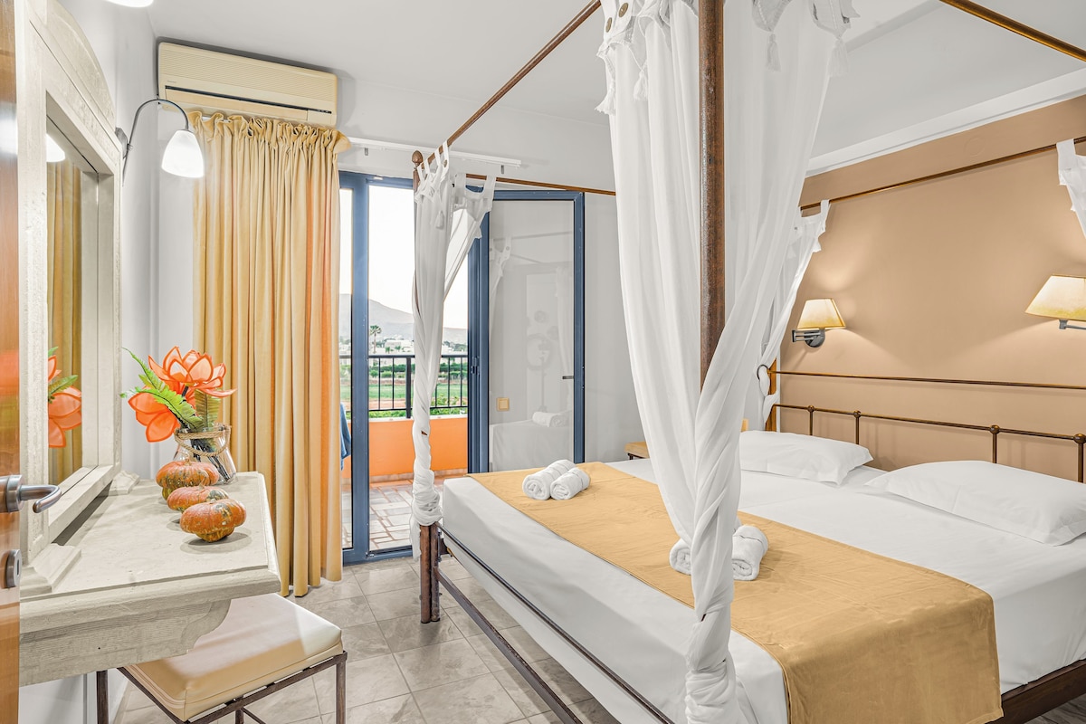 A hotel room with a bed, a bedside table, a mirror, a window with curtains, and a view of the outdoors.