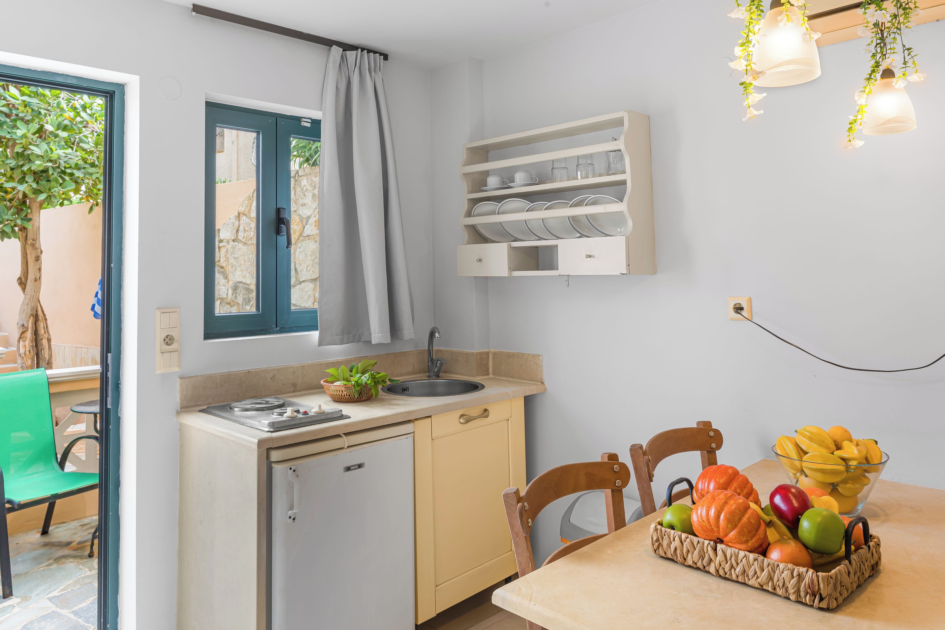 A kitchen with a sink, a small refrigerator, a window with a curtain, and a fruit basket on the table.