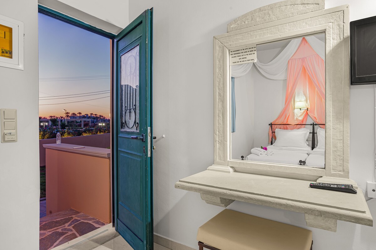 A hotel room with a large mirror, a bedside table, a bench, and a view of a cityscape through an open door.