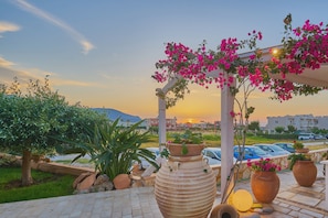 Ein gepflasterter Außenbereich mit großen Terrakottatöpfen, einer Pergola mit rosa blühenden Weinreben und einem Sonnenuntergang im Hintergrund.