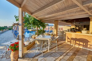 An outdoor patio with tables and chairs, a bar area, and a pool in the background. - Golden Bay Apartments-garden Haven (Hersonissos)