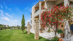 A hotel with a well-maintained lawn, stone pillars, and a balcony with red flowers. - Golden Bay Apartments-garden Haven (Hersonissos)