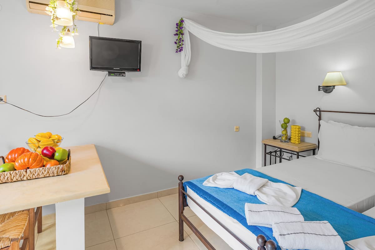 Habitación de hotel con cama, televisor, una mesa con fruta, lámpara y una mesita con planta.