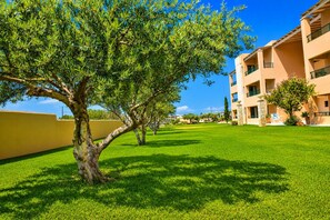 A well-maintained courtyard with a large tree, a building with balconies, and a clear blue sky. - Golden Bay Apartments-garden Haven (Hersonissos)