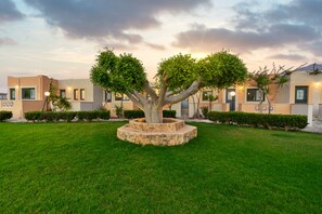 A tree in a stone planter surrounded by a well-maintained lawn, with buildings in the background. - Golden Bay Apartments-garden Haven (Hersonissos)