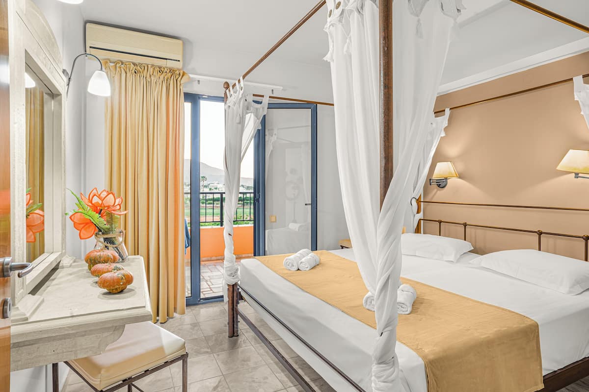 Habitación de hotel con cama, mesita de noche, espejo, ventana con cortinas y vistas al exterior.