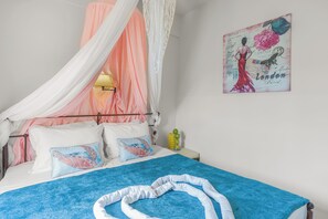 Un lit avec une couverture bleue, des oreillers blancs et un baldaquin blanc. Il y a un tableau accroché au mur et une petite table avec une lampe.